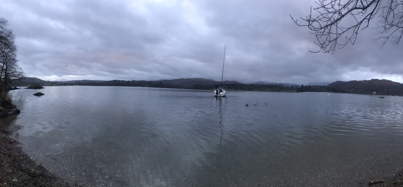 A lake under a grey sky, with a moored sailing boat and wooded hills beyond
